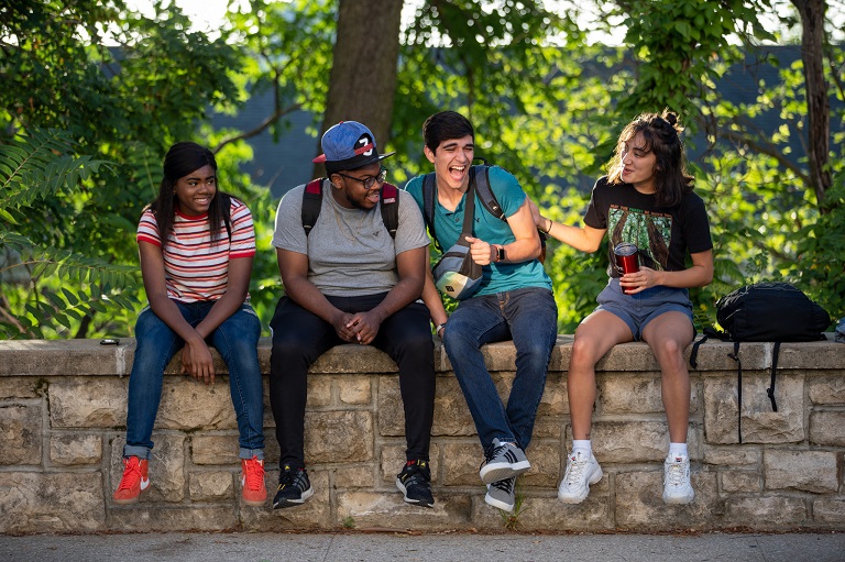Four students sit on a stone wall on the KU campus and are laughing.
